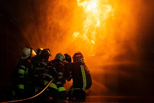 Bild vergr&ouml;&szlig;ern: Gruppe von Feuerwehrleuten, die über Wasserschlauch die Flammen in einer gefährlichen Brandstelle bei Nacht bekämpft.